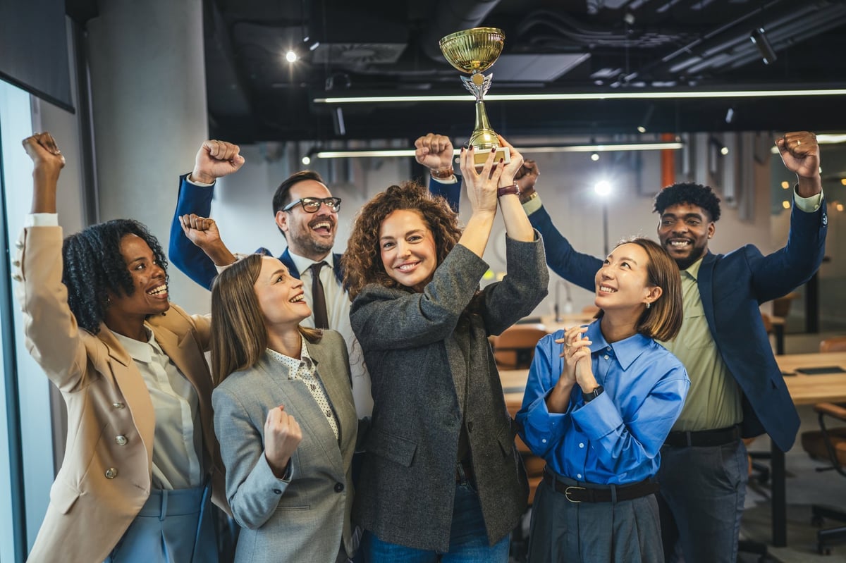 Diverse business people raising arms and cheering while a smiling woman holds a golden trophy, celebrating a major achievement and success in a modern office